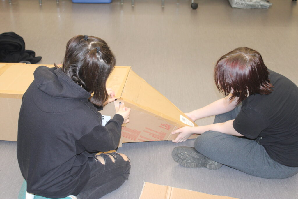 Two students sit on the floor and tape together a cardboard canoe. 