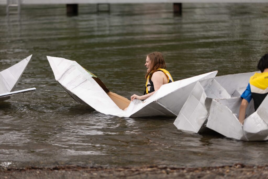 The author in a collapsing cardboard boat on a lake. She is laughing. 